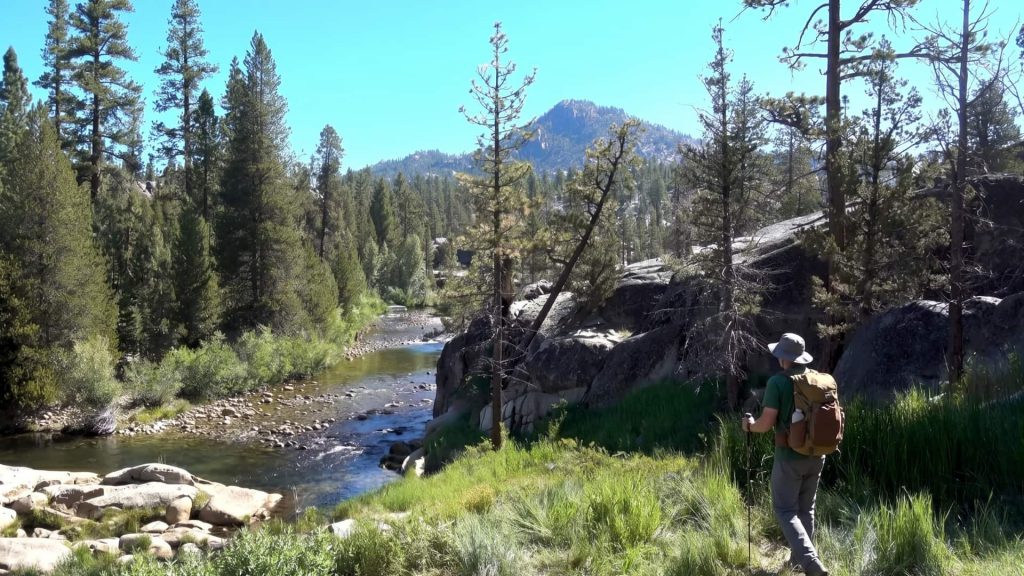 Hiker with backpack and trekking pole walking a riverside trail with granite slopes and Sierra peaks near Mono Hot Springs