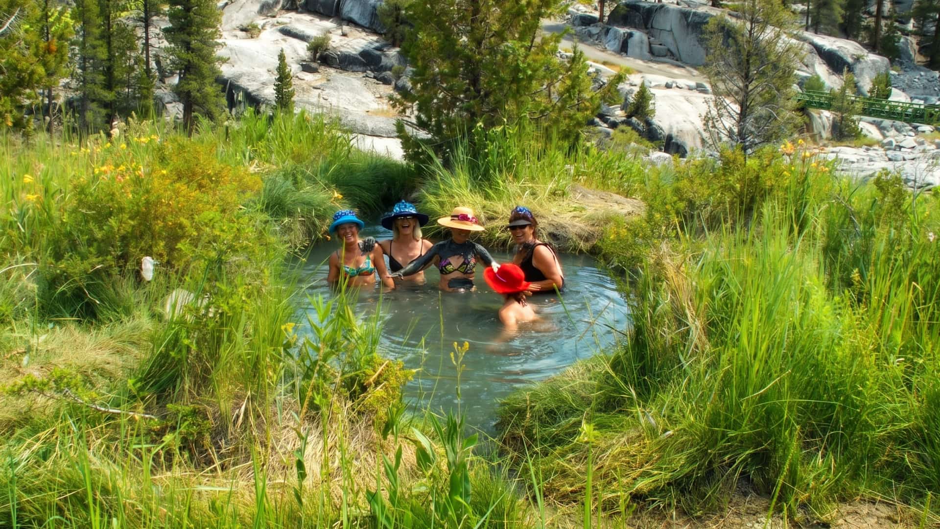 Group of friends in sun hats soaking in a natural mineral hot spring pool surrounded by tall grass, wildflowers, pine trees, and granite cliffs at Mono Hot Springs in the High Sierra