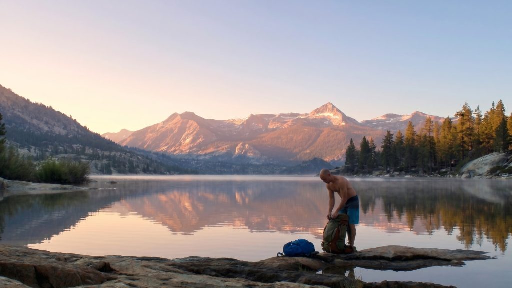 Hiker packing gear at a calm Florence lake with golden-hour mountain reflections in the Sierra Nevada near Mono Hot Springs Resort
