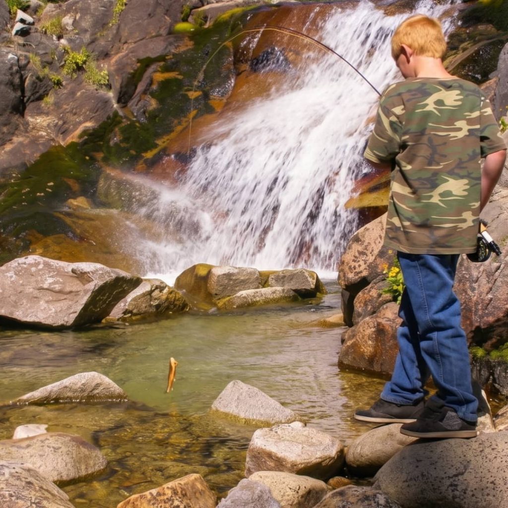 Young angler casting a fishing rod into a clear pool below a small waterfall surrounded by granite boulders and pine wildflowers near Mono Hot Springs