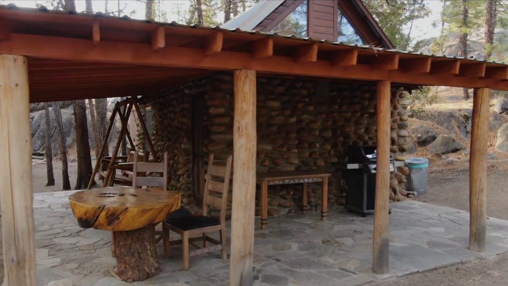 Covered stone patio of the Honeymoon Cabin with a wood-slab table, porch swing, and grill set among the pines